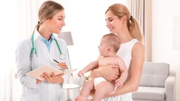 Médica conversando com uma mãe que segura um bebê, enquanto anota em uma prancheta, em uma consulta pediátrica para verificar sintomas de doenças comuns, como a faringite em bebês.