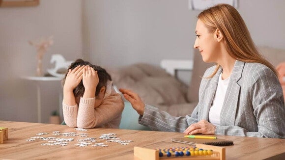 Uma criança pequena, com as mãos cobrindo o rosto, parece frustrada enquanto está sentada à mesa com um quebra-cabeça desmontado e um ábaco. Uma mulher adulta, com uma expressão gentil, coloca a mão no ombro da criança, tentando oferecer apoio. A cena captura um momento de desafio e a busca por compreensão durante a fase do não infantil. Uma criança pequena, com as mãos cobrindo o rosto, parece frustrada enquanto está sentada à mesa com um quebra-cabeça desmontado e um ábaco. Uma mulher adulta, com uma expressão gentil, coloca a mão no ombro da criança, tentando oferecer apoio. A cena captura um momento de desafio e a busca por compreensão durante a fase do não infantil.