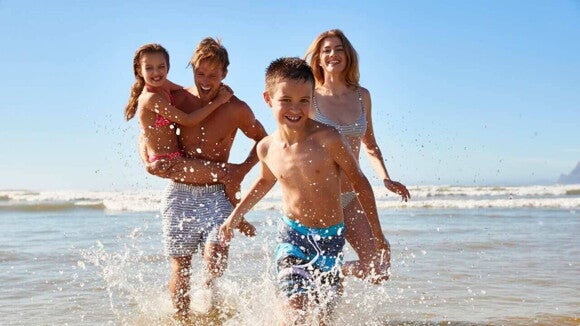 Família sorridente se divertindo na praia. O pai carrega a filha no colo, enquanto a mãe e o filho andam livremente pelas águas do mar Família sorridente se divertindo na praia. O pai carrega a filha no colo, enquanto a mãe e o filho andam livremente pelas águas do mar