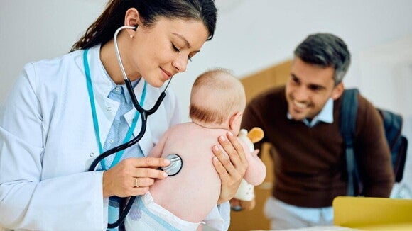 Uma médica, segurando um bebê nos braços e examinando ele com um estetoscópio. Ao fundo, um homem sorrindo para o bebê Uma médica, segurando um bebê nos braços e examinando ele com um estetoscópio. Ao fundo, um homem sorrindo para o bebê