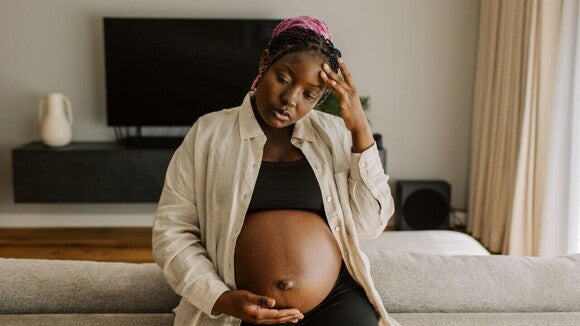 Mulher grávida com uma camisa bege, sentada com uma mão sobre a barriga e outra na testa, aparentando estar desconfortável. Mulher grávida com uma camisa bege, sentada com uma mão sobre a barriga e outra na testa, aparentando estar desconfortável.
