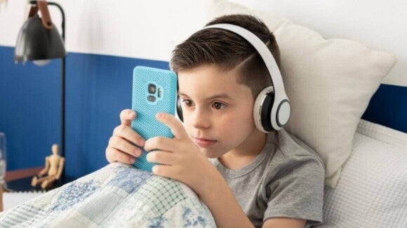 Criança concentrada no celular e utilizando fones de ouvido em seu quarto azul e branco. Criança concentrada no celular e utilizando fones de ouvido em seu quarto azul e branco.