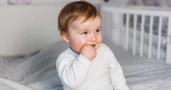 Menino pequeno de aproximadamente 1 ano sentado na cama com a mão na boca. Menino pequeno de aproximadamente 1 ano sentado na cama com a mão na boca.