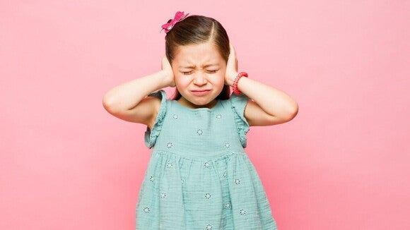 Uma menina de vestido azul expressando desconforto pela hipersensibilidade sensorial infantil. Ela está com os olhos fechados, a expressão franzida e as mãos pressionando firmemente os ouvidos contra um fundo rosa, indicando incômodo com ruídos altos.
