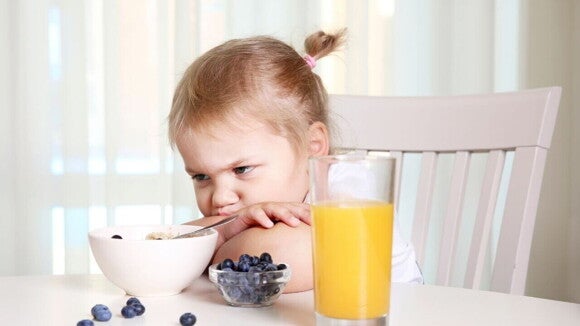Criança sentada em uma mesa com a cabeça deitada sobre os braços, com olhar irritado, observando um pote de comida à sua frente. Criança sentada em uma mesa com a cabeça deitada sobre os braços, com olhar irritado, observando um pote de comida à sua frente.