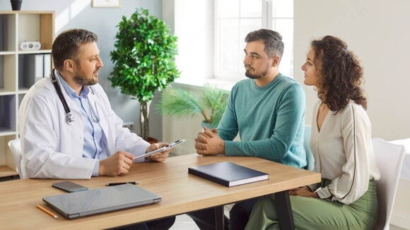 Homem e mulher sentados à mesa, conversando seriamente com um médico em seu consultório. O médico está com um estetoscópio e segurando uma prancheta.