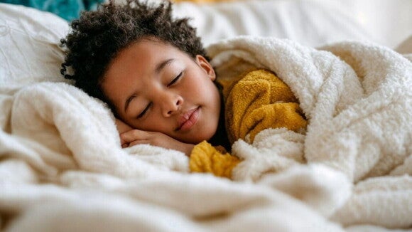Uma criança dormindo profundamente de lado na cama, aconchegada sob um cobertor branco e vestindo uma blusa amarela.