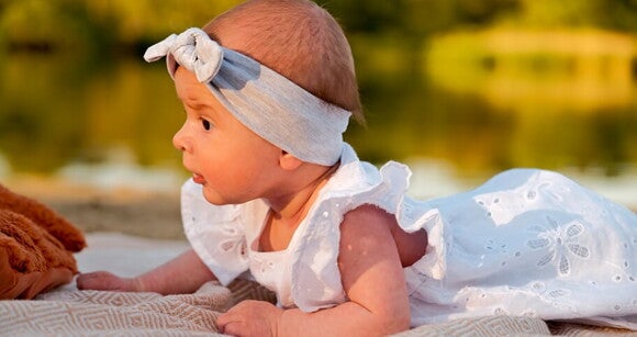Bebê deitada de bruços usando faixa na cabeça e vestido branco Bebê deitada de bruços usando faixa na cabeça e vestido branco