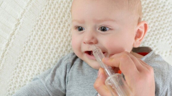 Um bebê deitado com a boca aberta, enquanto uma mão segura uma seringa próxima ao nariz do bebê