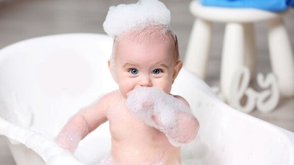 Um bebê está sentado em uma banheira cheia de espuma. O bebê tem espuma na cabeça e nos braços, com uma expressão sorridente.