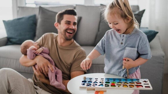 Um homem sorrindo e sentado no chão com um bebê no colo, ao lado de uma criança em pé e sorrindo, brincando com objetos sobre uma mesa. Um homem sorrindo e sentado no chão com um bebê no colo, ao lado de uma criança em pé e sorrindo, brincando com objetos sobre uma mesa.