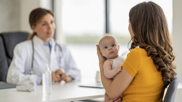 Uma mulher segurando um bebê nos braços, enquanto está sentada em uma mesa, em frente a uma profissional da saúde. Uma mulher segurando um bebê nos braços, enquanto está sentada em uma mesa, em frente a uma profissional da saúde.