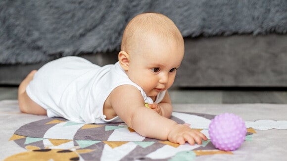 Um bebê deitado de bruços em um tapete, estendendo a mão para alcançar uma bola de brinquedo texturizada, demonstrando um marco de desenvolvimento.