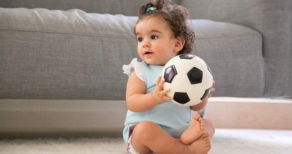 Bebê sentada em um tapete e segurando uma bola de futebol Bebê sentada em um tapete e segurando uma bola de futebol