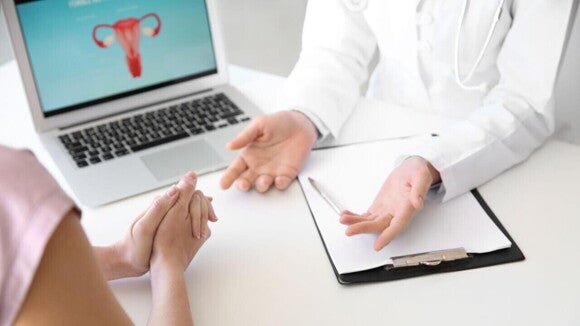 Mãos de uma paciente e um médico sob uma mesa de consultório, ao lado de um computador com a ilustração de um útero Mãos de uma paciente e um médico sob uma mesa de consultório, ao lado de um computador com a ilustração de um útero