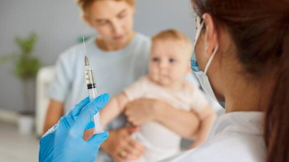 Uma profissional de saúde, de máscara e luvas azuis, segura uma seringa com a dose da vacina, pronta para a aplicação. No fundo, desfocado, uma mãe segura um bebê no colo, preparando-o para receber a vacina contra a meningite B.