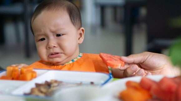 Um bebê visivelmente chateado e choroso, usando um babador, está sentado em uma cadeira de alimentação e um adulto está oferecendo um pedaço de melancia.