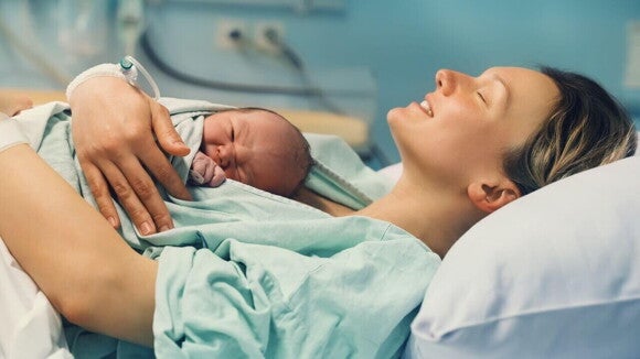 Mulher deitada em uma maca, com semblante de satisfação, segurando um recém-nascido nos braços. Mulher deitada em uma maca, com semblante de satisfação, segurando um recém-nascido nos braços.