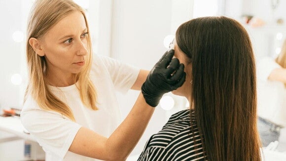 Uma mulher com as mãos enluvadas, está realizando um procedimento facial em outra mulher, que está de costas para o observador. Uma mulher com as mãos enluvadas, está realizando um procedimento facial em outra mulher, que está de costas para o observador.