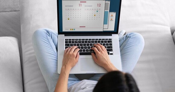 Mulher sentada em um sofá com um notebook aberto em seu colo olhando para um calendário Mulher sentada em um sofá com um notebook aberto em seu colo olhando para um calendário