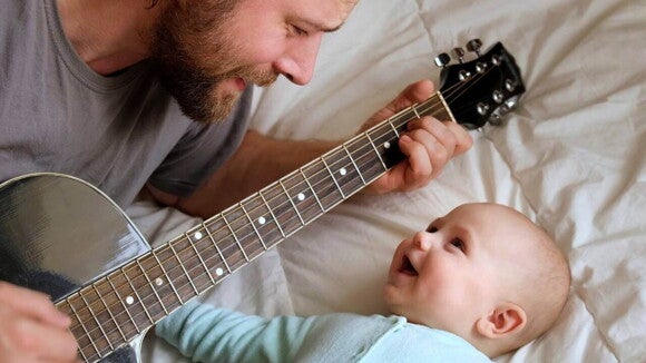 Música para bebês Close-up de um pai tocando um violão enquanto olha carinhosamente para seu bebê deitado ao lado dele, em uma cama.