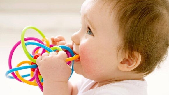 Nascimento dos dentes bebê Bebê segurando e mordendo um brinquedo colorido de plástico, em fase oral, indicando o início do nascimento dos dentes do bebê.