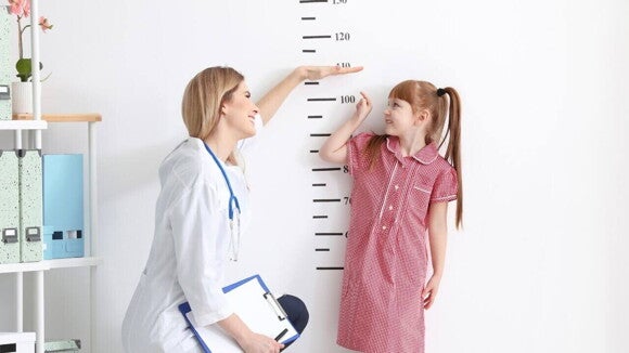 Uma profissional de saúde e uma menina, apontando para uma marcação de altura em um consultório médico. Uma profissional de saúde e uma menina, apontando para uma marcação de altura em um consultório médico.