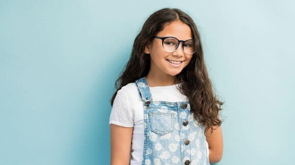 Menina de cabelos pretos e longos usando óculos e macacão jeans em frente a uma parede azul claro. Menina de cabelos pretos e longos usando óculos e macacão jeans em frente a uma parede azul claro.