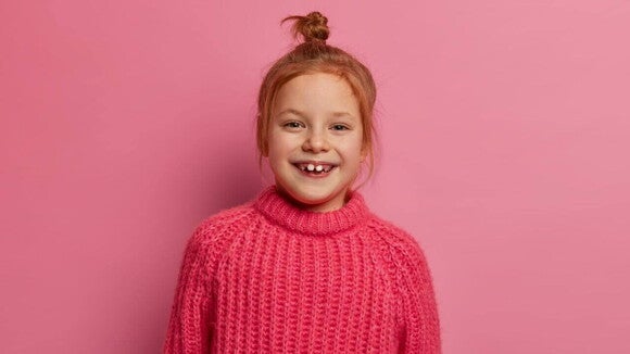 Uma menina de aproximadamente 6 anos sorrindo, com um suéter rosa, de frente para uma parede rosa. Uma menina de aproximadamente 6 anos sorrindo, com um suéter rosa, de frente para uma parede rosa.