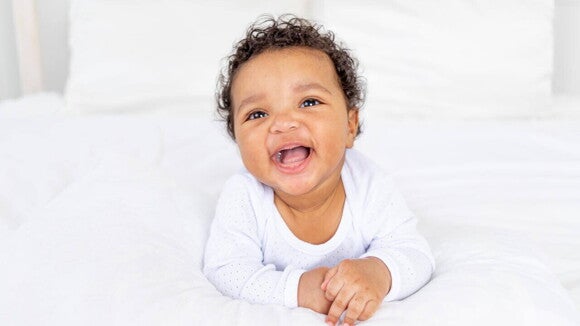 Um bebê de aproximadamente 6 meses sorrindo e deitado em uma cama, usando um body branco. Um bebê de aproximadamente 6 meses sorrindo e deitado em uma cama, usando um body branco.