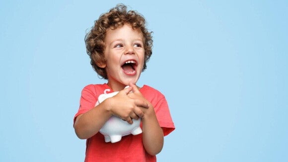Menino sorridente com os cabelos cacheados abraçando um cofrinho branco em um fundo azul. Menino sorridente com os cabelos cacheados abraçando um cofrinho branco em um fundo azul.