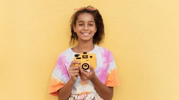 Menina segurando câmera fotográfica e sorrindo em frente a uma parede amarela. Menina segurando câmera fotográfica e sorrindo em frente a uma parede amarela.