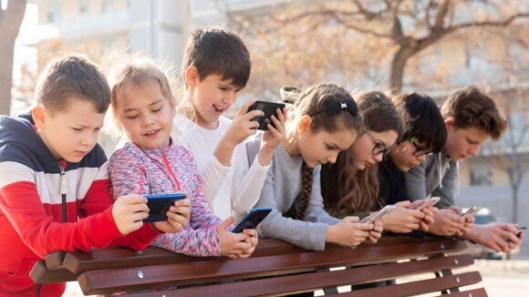 Um grupo de crianças inclina-se sobre um banco de parque e todas estão focadas nos seus celulares. Um grupo de crianças inclina-se sobre um banco de parque e todas estão focadas nos seus celulares.