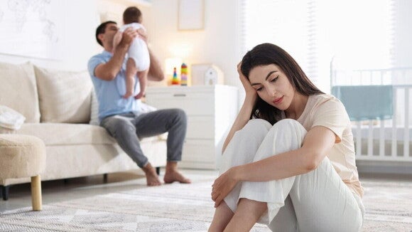Uma mãe, vestindo roupas claras, está sentada no chão, abraçando os joelhos e olhando para baixo com expressão de tristeza ou melancolia, indicando um possível baby blues ou depressão pós-parto. Ao fundo, desfocado, o pai está sentado em um sofá, brincando com o bebê.