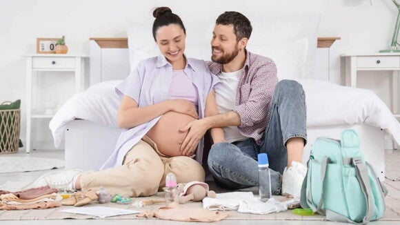 Casal grávido sorrindo, sentados no chão do quarto e cercados por itens de bebê, enquanto tocam a barriga. A cena mostra a preparação para a chegada do bebê e destaca a questão do que levar na bolsa maternidade, organizando os itens essenciais. Casal grávido sorrindo, sentados no chão do quarto e cercados por itens de bebê, enquanto tocam a barriga. A cena mostra a preparação para a chegada do bebê e destaca a questão do que levar na bolsa maternidade, organizando os itens essenciais.