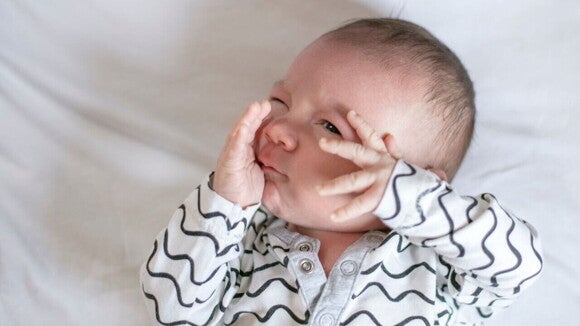 Um bebê deitado com as duas mãos no rosto, e um dos olhos fechados Um bebê deitado com as duas mãos no rosto, e um dos olhos fechados