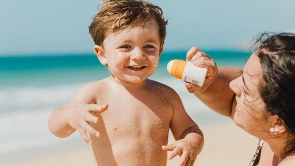 Uma criança na praia sorrindo, enquanto uma mulher passa protetor solar no seu rosto Uma criança na praia sorrindo, enquanto uma mulher passa protetor solar no seu rosto