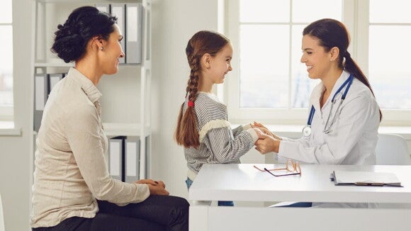 Uma médica sorridente está sentada e segurando as mãos de uma menina que também sorri, enquanto sua mãe, sentada à esquerda, também sorri. Uma médica sorridente está sentada e segurando as mãos de uma menina que também sorri, enquanto sua mãe, sentada à esquerda, também sorri.