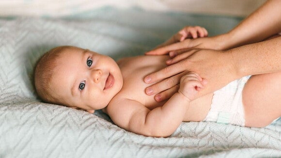 Um bebê sorridente deitado de costas em um cobertor, enquanto as mãos de um adulto o tocam gentilmente nas laterais do corpo. Um bebê sorridente deitado de costas em um cobertor, enquanto as mãos de um adulto o tocam gentilmente nas laterais do corpo.