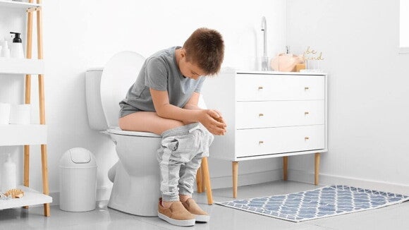 Menino sentado no vaso sanitário com as calças abaixadas, em um banheiro claro. A imagem representa a dificuldade e o tempo gasto para evacuar, um sintoma comum da prisão de ventre infantil. Menino sentado no vaso sanitário com as calças abaixadas, em um banheiro claro. A imagem representa a dificuldade e o tempo gasto para evacuar, um sintoma comum da prisão de ventre infantil.