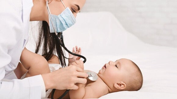 Pediatra com máscara examinando um bebê deitado em uma maca branca com um estetoscópio.
