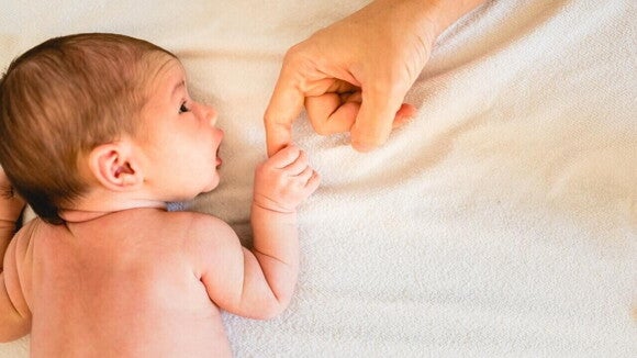 Close-up de um recém-nascido deitado em um cobertor branco segurando o dedo de um adulto. A imagem representa o cuidado e o monitoramento da saúde do bebê após a avaliação de qual o peso normal de um recém-nascido.