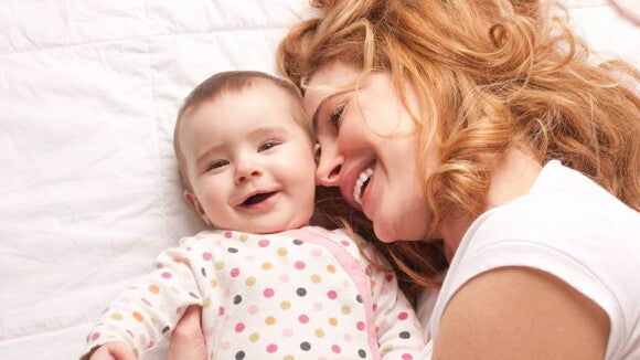 Uma mãe está deitada ao lado de seu filho no berço. Ambos sorriem alegremente para a câmera. O bebê veste um macacão branco com bolinhas coloridas.