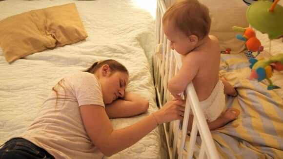 Mãe deitada ao lado do berço enquanto o bebê acordado a observa, ilustrando os desafios da regressão do sono dos 2 anos e o cansaço da família.