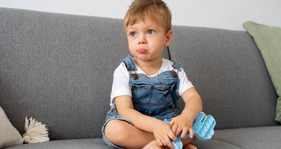 Menino usando macacão jeans sentado no sofá segurando peças de um brinquedo de montar. Menino usando macacão jeans sentado no sofá segurando peças de um brinquedo de montar.