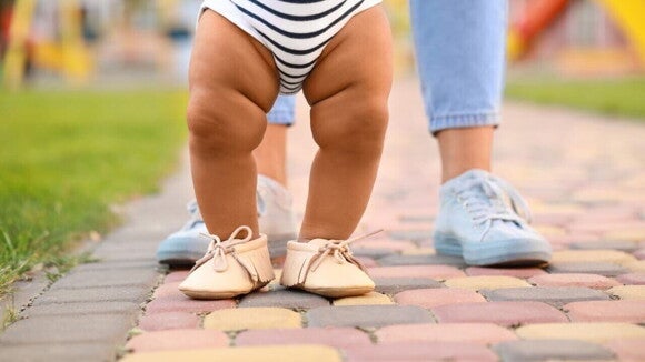 Pernas de um bebê calçando um sapato e andando sobre um chão de pedras, com o apoio de um adulto. Pernas de um bebê calçando um sapato e andando sobre um chão de pedras, com o apoio de um adulto.