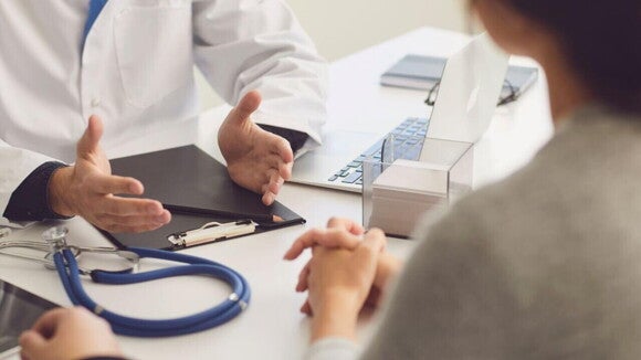 Médico de jaleco em uma consulta, gesticulando com as mãos enquanto conversa com um paciente, discutindo o diagnóstico e tratamento de condições de pele, como a sarna infantil.