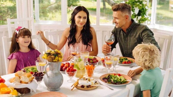 Uma família de quatro pessoas, incluindo um casal adulto e duas crianças (uma menina e um menino), está sentada à mesa em um ambiente claro e arejado.. A mesa está posta com uma variedade de alimentos saudáveis como saladas, frutas, pães e sucos. A imagem contrasta com a seletividade alimentar, mostrando uma refeição familiar harmoniosa.