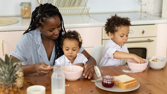 Uma mãe e seus dois filhos pequenos estão sentados em uma mesa de café da manhã. A mãe, sorrindo, ajuda a bebê a se alimentar, enquanto, ao lado, um menino mais velho, brinca com a comida em sua própria tigela.