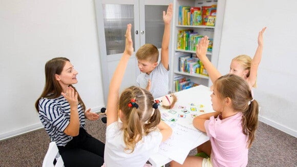 Grupo de crianças sentadas em uma mesa com a mão para o alto, ao lado de uma mulher. Grupo de crianças sentadas em uma mesa com a mão para o alto, ao lado de uma mulher.